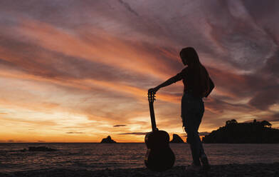 Silhouette einer Frau mit Gitarre, die bei Sonnenuntergang am Strand steht, Almunecar, Spanien - LJF01216