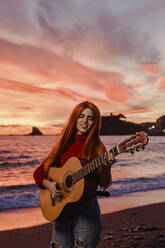 Porträt einer rothaarigen jungen Frau, die bei Sonnenuntergang am Strand Gitarre spielt, Almunecar, Spanien - LJF01219