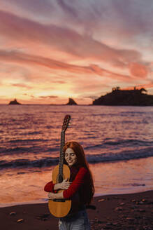 Porträt einer rothaarigen jungen Frau, die am Strand bei Sonnenuntergang eine Gitarre umarmt, Almunecar, Spanien - LJF01222