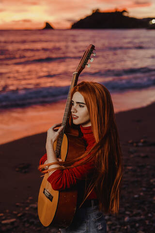 Porträt einer rothaarigen jungen Frau mit Gitarre am Strand bei Sonnenuntergang mit Blick in die Ferne, Almunecar, Spanien, lizenzfreies Stockfoto