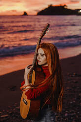 Portrait of redheaded young woman with guitar on the beach at sunset looking at distance, Almunecar, Spain - LJF01223