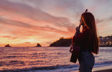 Junge Frau mit Gitarre, die bei Sonnenuntergang am Strand steht und auf das Meer schaut, Almunecar, Spanien - LJF01224