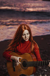 Porträt einer rothaarigen jungen Frau, die bei Sonnenuntergang am Strand Gitarre spielt, Almunecar, Spanien - LJF01225
