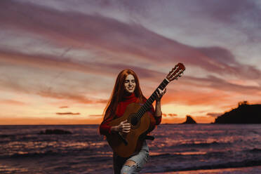 Portrait of redheaded young woman playing guitar on the beach at sunset, Almunecar, Spain - LJF01226