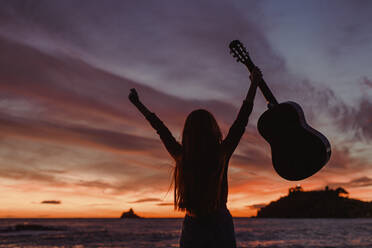 Silhouette einer Frau mit Gitarre, die bei Sonnenuntergang am Strand steht, Almunecar, Spanien - LJF01227