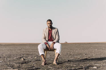 Portrait of young man sitting on chair in barren land - ERRF02444