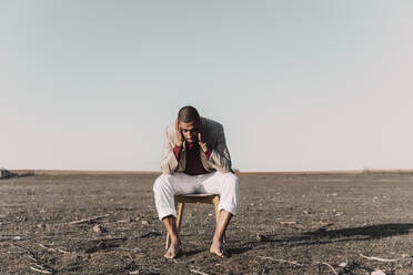 Desperate young man sitting on chair in barren land - ERRF02445