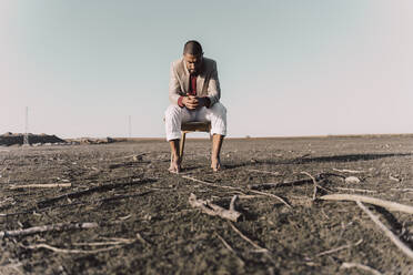 Pensive young man sitting on chair in barren land - ERRF02447