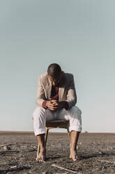 Pensive young man sitting on chair in barren land - ERRF02449