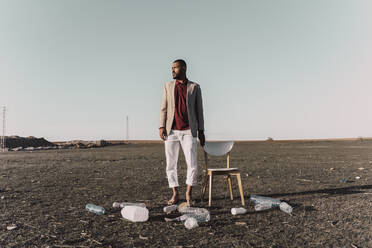 Junger Mann steht neben einem Stuhl, umgeben von Plastikflaschen in einem kargen Land - ERRF02459