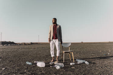 Young man standing next to chair surrounded by plastic bottles in barren land - ERRF02459