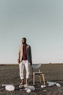 Young man standing next to chair surrounded by plastic bottles in barren land - ERRF02460