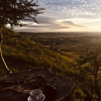 Tansania, Mount Meru Region, Blick auf den Kilimandscharo bei Sonnenaufgang mit Kaffeetasse auf dem Tisch im Vordergrund - MWEF00216