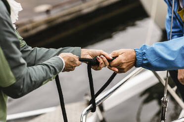 Cropped hands of female instructor and senior man tying rope on yacht's railing - MASF16000