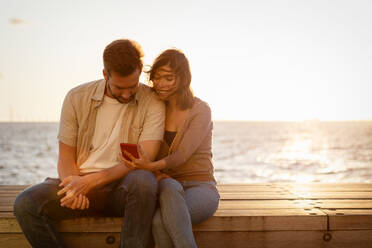 Lächelnde Frau, die ihrem Freund das Handy zeigt, während sie bei Sonnenuntergang auf einem Sitz am Meer sitzt - MASF16006