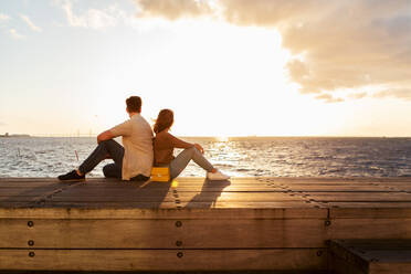 Paar in voller Länge auf einem Sitz am Meer sitzend gegen den Himmel bei Sonnenuntergang - MASF16008