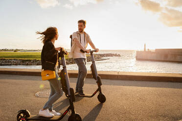 Couple talking while riding electric push scooters on road by sea against sky - MASF16011