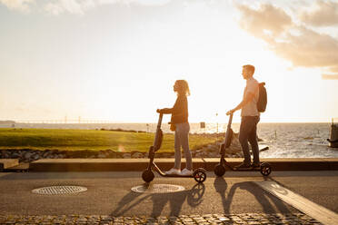Full length of couple enjoying on electric push scooters by sea against sky during sunset - MASF16012