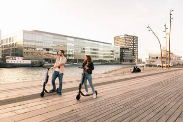 Couple exploring city while riding on electric push scooters at promenade by river - MASF16016