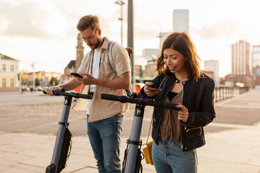 Male and female commuters scanning through mobile phone while unlocking electric push scooters in city - MASF16018