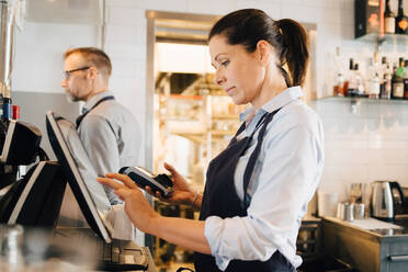 Der Besitzer benutzt einen Computer, während er ein Kreditkartenlesegerät in einem Restaurant hält - MASF16055