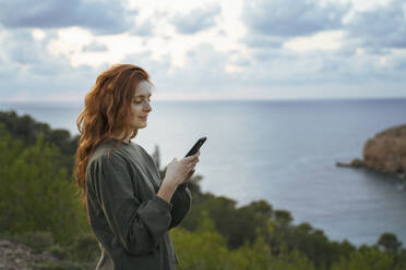 Rothaarige junge Frau, die an der Küste ein Mobiltelefon benutzt, Ibiza, Spanien - AFVF04835