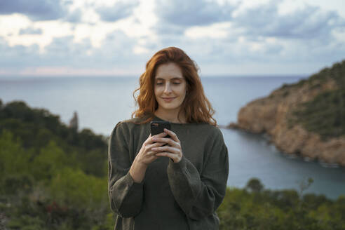 Rothaarige junge Frau, die an der Küste ein Mobiltelefon benutzt, Ibiza, Spanien - AFVF04836