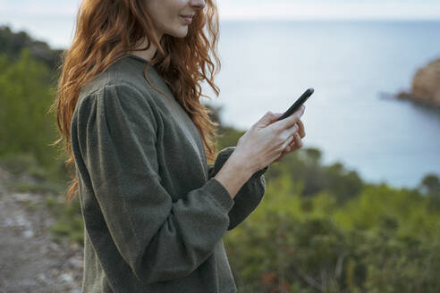 Rothaarige junge Frau, die an der Küste ein Mobiltelefon benutzt, Ibiza, Spanien - AFVF04838