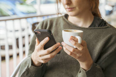 Nahaufnahme einer jungen Frau, die ein Mobiltelefon benutzt und eine Kaffeetasse hält - AFVF04839