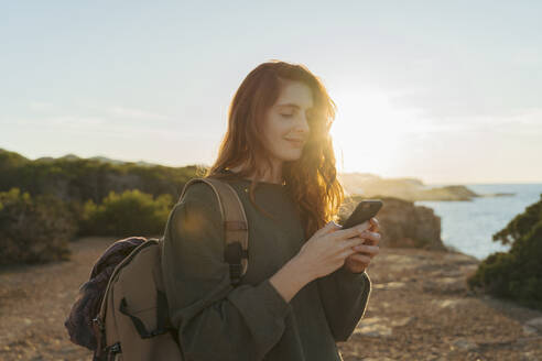 Rothaarige junge Frau mit Handy an der Küste bei Sonnenuntergang, Ibiza, Spanien - AFVF04858