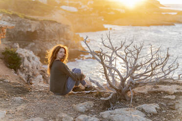 Rothaarige junge Frau, die bei Sonnenuntergang an der Küste sitzt, Ibiza, Spanien - AFVF04861