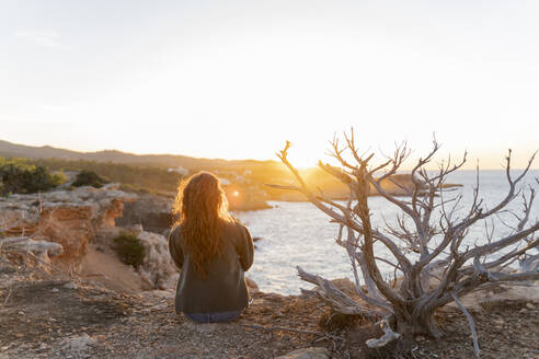 Rothaarige junge Frau, die bei Sonnenuntergang an der Küste sitzt, Ibiza, Spanien - AFVF04863