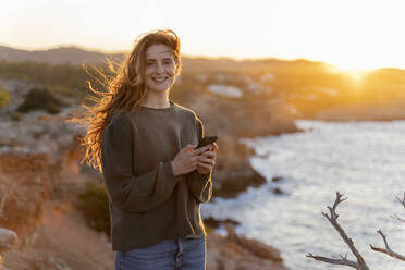 Porträt einer lächelnden rothaarigen jungen Frau mit Mobiltelefon an der Küste bei Sonnenuntergang, Ibiza, Spanien - AFVF04865