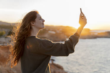 Rothaarige junge Frau macht ein Selfie an der Küste bei Sonnenuntergang, Ibiza, Spanien - AFVF04868