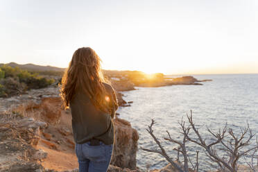 Rothaarige junge Frau, die bei Sonnenuntergang an der Küste steht, Ibiza, Spanien - AFVF04869
