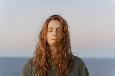 Rothaarige junge Frau mit geschlossenen Augen an der Küste bei Sonnenuntergang, Ibiza, Spanien - AFVF04872