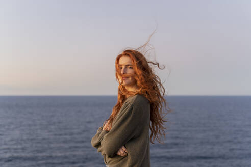 Porträt einer rothaarigen jungen Frau mit vom Wind zerzausten Haaren an der Küste bei Sonnenuntergang, Ibiza, Spanien - AFVF04874