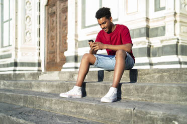 Junger Mann sitzt auf einer Außentreppe und überprüft sein Smartphone, Florenz, Italien - FBAF01138