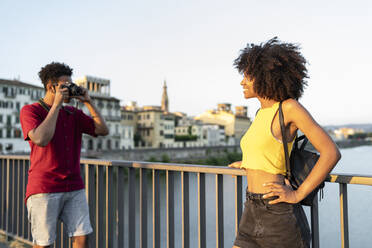 Junger Mann fotografiert seine Freundin auf einer Brücke über dem Fluss Arno bei Sonnenuntergang, Florenz, Italien - FBAF01160