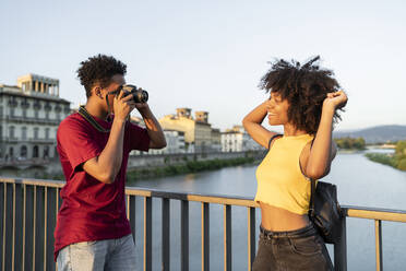 Junger Mann fotografiert seine Freundin auf einer Brücke über dem Fluss Arno bei Sonnenuntergang, Florenz, Italien - FBAF01161