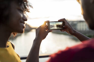 Junges Touristenpaar beim Fotografieren auf einer Brücke über dem Fluss Arno bei Sonnenuntergang, Florenz, Italien - FBAF01167