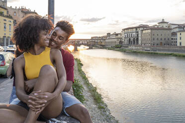 Verliebtes junges Touristenpaar sitzt auf einer Mauer am Fluss Arno bei Sonnenuntergang, Florenz, Italien - FBAF01169