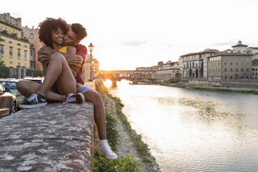 Verliebtes junges Touristenpaar sitzt auf einer Mauer am Fluss Arno bei Sonnenuntergang, Florenz, Italien - FBAF01170