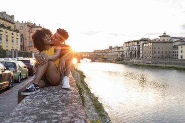 Verliebtes junges Touristenpaar sitzt auf einer Mauer am Fluss Arno bei Sonnenuntergang, Florenz, Italien - FBAF01171