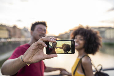 Young tourist couple taking a selfie on a bridge above river Arno at sunset, Florence, Italy - FBAF01183