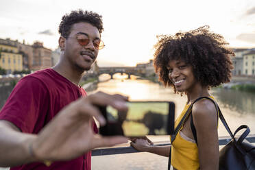 Young tourist couple taking a selfie on a bridge above river Arno at sunset, Florence, Italy - FBAF01184