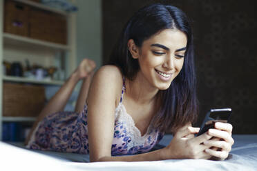 Portrait of smiling young woman lying on bed looking at smartphone - JSMF01428