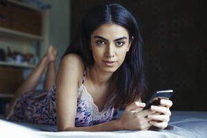 Porträt einer jungen Frau auf dem Bett liegend mit Smartphone - JSMF01429