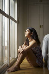 Young woman sitting on the floor of her bedroom enjoying sunlight in the morning - JSMF01431