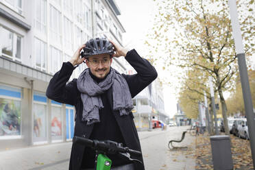 Porträt eines lächelnden jungen Mannes, der mit einem Elektroroller auf dem Bürgersteig steht und einen Fahrradhelm aufsetzt - KMKF01152